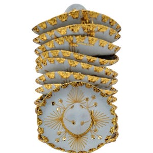 May include: A stack of white miniature sombreros featuring gold metallic trim along the brims and intricate gold thread embroidery on the crowns. The hats are arranged vertically, showcasing the consistent circular design and reflective gold accents.