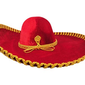 Op de afbeelding: Een rode fluwelen sombrero met een brede rand en een hoge, ronde kroon. De hoed is versierd met een gouden gevlochten bies langs de rand van de hoed en rond de basis van de kroon, inclusief een decoratieve gouden knoop en een hangend koord.