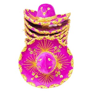 Puede incluir: Una pila de sombreros de color fucsia brillante con ribete metálico dorado a lo largo del ala ancha y la copa. Cada sombrero está decorado con bordados de hilo dorado en un patrón de rayos de sol y pequeñas lentejuelas doradas circulares.