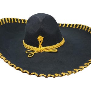 May include: A black sombrero featuring a wide brim with gold-coloured braided trim along the edge. The crown is adorned with a matching gold-coloured braided cord that wraps around the base and ties into a decorative knot at the front centre.
