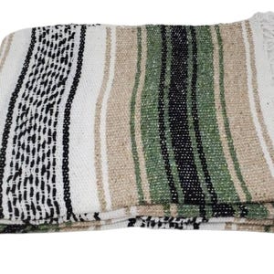 May include: A woven blanket featuring a striped pattern in shades of white, black, and tan with green accents. The textile has a textured weave and is finished with white fringe along the short edge, displaying a repeating geometric design.