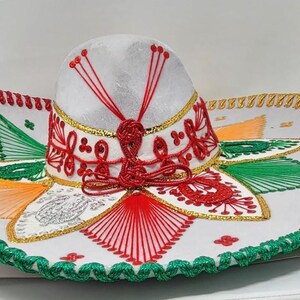 May include: A white sombrero with intricate embroidery in red, green, gold, and orange. The hat features a rounded crown and a wide, decorated brim. The brim is trimmed with green and gold stitching, and the crown has red accents.