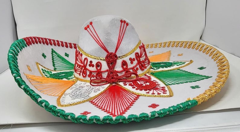 May include: A white sombrero with intricate embroidery in red, green, gold, and orange. The hat features a rounded crown and a wide, decorated brim. The brim is trimmed with green and gold stitching, and the crown has red accents.