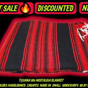 Puede incluir: Una manta plegada de nostalgia de Tijuana de los años 80 con rayas verticales rojas y negras. La manta tejida a mano tiene flecos blancos y mide aproximadamente 203 x 150 cm. La imagen incluye el texto "HOT SALE", "DISCOUNTED" y "NEW".