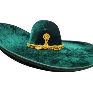 May include: A dark green velvet sombrero featuring a wide, upturned brim and a high, rounded crown. The base of the crown is adorned with a decorative gold braided trim that forms a knot detail at the front, adding texture to the smooth fabric.