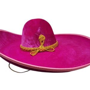 May include: A vibrant fuchsia sombrero featuring a soft velvet-like texture, a wide brim with a light pink piped edge, and a decorative gold braided trim encircling the base of the crown, complete with a gold braided bow detail at the front.