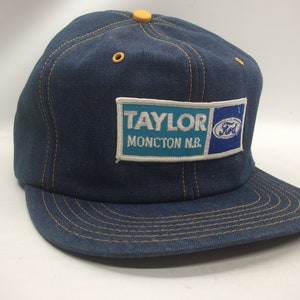 May include: A blue denim baseball cap with a yellow stitched brim. The cap has a patch on the front that says "Taylor Moncton N.B." and a Ford logo.