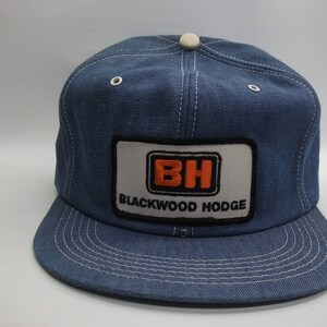 May include: A denim baseball cap with a flat brim and a white button on top. The cap features a rectangular patch with the letters "BH" in orange and the words "BLACKWOOD HODGE" in black.
