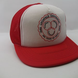 May include: Red and white trucker hat with a white patch featuring a circular logo that says "1886 Litchfield Centennial 1986 100 Years of Unity" with three overlapping circles that say "Families", "Neighbors", and "Communities".