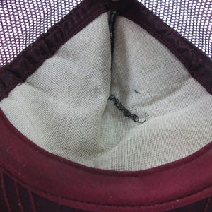 May include: The inside of a burgundy mesh trucker hat. The hat has a white cotton lining and a burgundy brim.