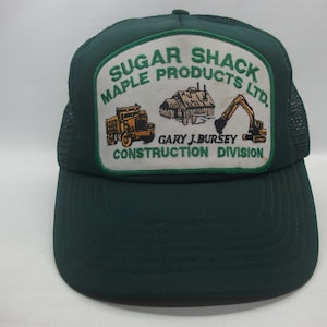 Peut inclure: Casquette de camionneur verte avec un patch blanc qui dit "Sugar Shack Maple Products Ltd. Gary J. Bursey Construction Division". Le patch présente également un camion à benne basculante, un bâtiment et une excavatrice.