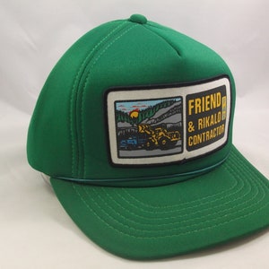 May include: Green trucker hat with a yellow and black patch that says "Friend & Rikalo Contractor" and a smaller patch with a picture of a construction site.