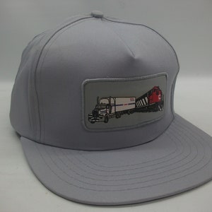 Peut inclure: Un chapeau de camionneur gris avec un patch représentant un semi-remorque blanc tirant une remorque blanche avec un logo CN rouge et blanc, suivi d'une locomotive noire et rouge.