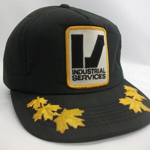 May include: Black trucker hat with a yellow and white patch that reads "Industrial Services". The hat has a gold embroidered maple leaf design around the brim.