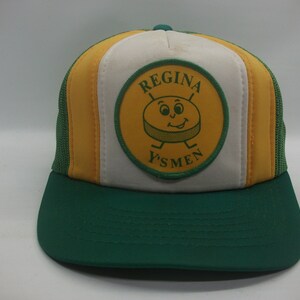 May include: A green, white, and yellow trucker hat with a patch that says "Regina Y's Men" and features a cartoon puck with a smiling face.