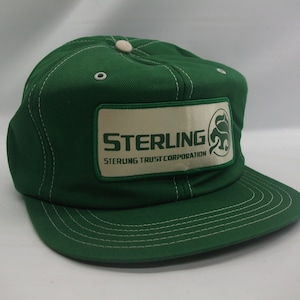 May include: Green baseball cap with a white patch that says "STERLING STERLING TRUST CORPORATION" with a green logo of a stylized animal.