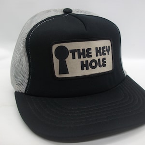 May include: A black and gray trucker hat with a white mesh back. The hat has a patch on the front that says "The Key Hole" with a keyhole graphic.