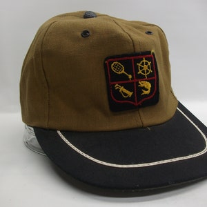 May include: A brown baseball cap with a black brim and a white stitching detail. The cap has a patch with a shield design featuring a tennis racket, a ship's wheel, a fishing rod, and a fish.