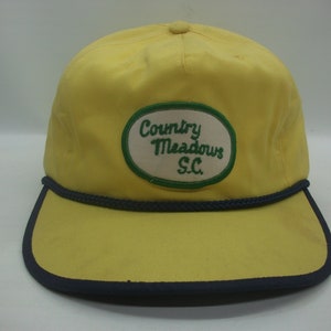 May include: Yellow baseball cap with a navy blue brim and a green and white oval patch that reads "Country Meadows S.C."