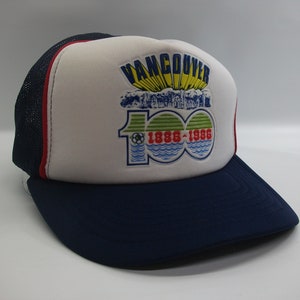 May include: A white and blue trucker hat with a red stripe. The front of the hat has a colorful embroidered patch that says "Vancouver 100 1886-1986" with a graphic of a city skyline and a soccer ball.