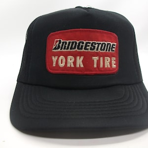 May include: Black trucker cap with a mesh back and a curved peak. It has a red rectangular patch on the front with the Bridgestone and York Tire logos in white. A classic style, suitable for casual wear.