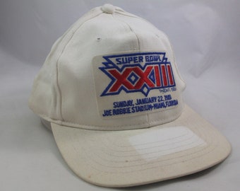 May include: White baseball cap with a blue and red Super Bowl XXIII patch. The patch includes the text "Super Bowl XXIII" and "January 22, 1989" and "Joe Robbie Stadium - Miami, Florida".