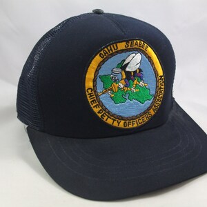 May include: A navy blue trucker hat with a yellow and green embroidered patch that says "Oahu Seabee Chief Petty Officers Association". The patch features a cartoon bee holding a wrench.