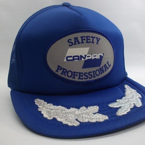 Peut inclure: Casquette de camionneur bleue avec un patch ovale blanc et gris qui dit "Safety Can-Par Professional". La casquette a deux feuilles de chêne brodées argentées sur la visière.