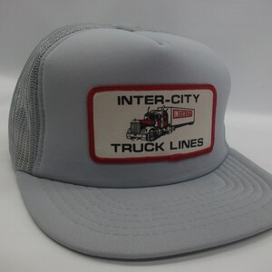 May include: A gray trucker hat with a red and white patch that says "Inter-City Truck Lines" and features a cartoon image of a semi-truck.