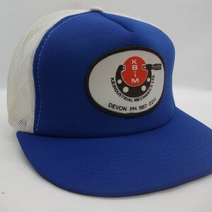 Peut inclure: Casquette de camionneur bleu et blanche avec un logo rouge, blanc et noir qui dit "K.B.I.M. Industrial Mechanics Ltd. Devon Ph. 987-2211"