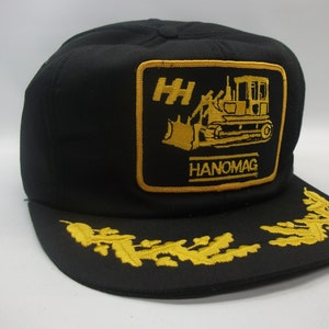 May include: Black trucker hat with a gold embroidered patch featuring a bulldozer and the text "HH HANOMAG". The hat also has a gold embroidered wreath around the brim.