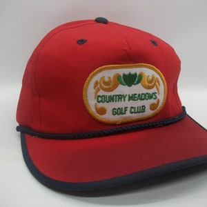 May include: Red baseball cap with a navy blue brim and a rope trim. The cap has a white oval patch with gold trim and green leaves. The patch reads "Country Meadows Golf Club".
