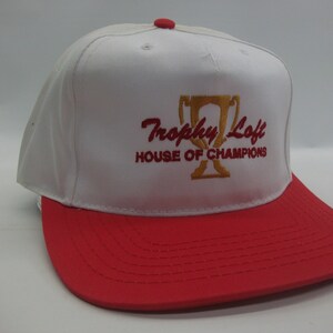May include: White and red baseball cap with the embroidered text "Trophy Loft House of Champions" and a gold trophy graphic.