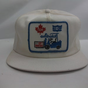 May include: White trucker hat with a blue and red patch featuring a forklift, the text "TCM", "BLU-CHIP", "AMHOIST", and "LIFT TRUCKS", and a Canadian maple leaf.