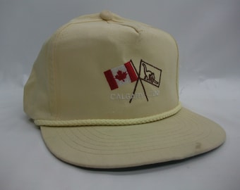 May include: A beige baseball cap with a white rope trim. The front of the cap features an embroidered design of a Canadian flag and a dinosaur flag crossed over each other. The text "CALGARY ZOO" is embroidered below the flags.