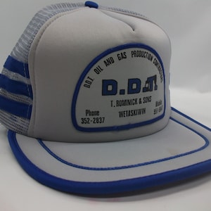 May include: Gray and blue trucker hat with a patch that reads "D.D.D.T. Oil and Gas Production Contractors T. Romnick & Sons Wetaskiwin Phone 352-2837 Mobile 551-5847"