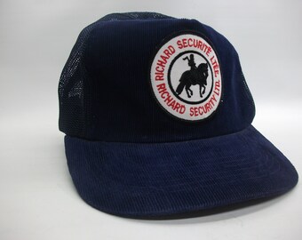 May include: A navy blue mesh trucker hat with a large, round patch featuring a white silhouette of a person riding a horse. The patch has the text "Richard Securite Ltee. Richard Security Ltd." in red and white.