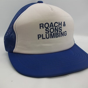 Puede incluir: Una gorra de camionero blanca y azul con el texto "ROACH & SONS PLUMBING" en letras azules.