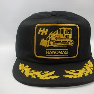 May include: Black trucker hat with a gold embroidered patch featuring a yellow bulldozer and the text "HH HANOMAG". The hat also has gold embroidered oak leaves on the brim.