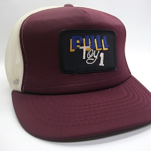 May include: A maroon trucker hat with a white mesh back. The hat has a black patch with the text "BULL Toy 1" in yellow, blue, and white lettering.