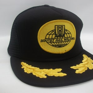 May include: Black trucker hat with a yellow embroidered patch that says "Boutet Rail Welding" and a globe with a building inside. The hat also has two yellow embroidered oak leaves on the brim.