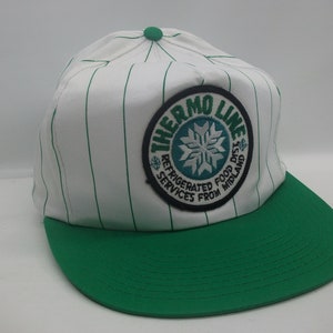 May include: White and green baseball cap with a green and white striped pattern. The cap has a circular patch with the text "Thermo Line" and a snowflake design. The text on the patch reads "Refrigerated Food Dist. Services From Midland."