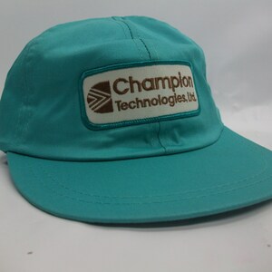 Peut inclure: Une casquette de baseball bleu turquoise avec un patch blanc sur lequel est inscrit en brun "Champion Technologies, Ltd."
