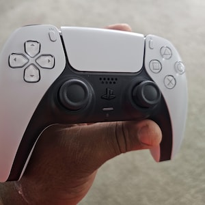 May include: A white and black wireless game controller with a central touchpad, directional buttons, and analog sticks. The controller has a sleek design with a PlayStation logo and various function buttons.