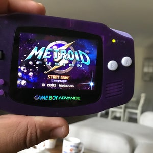 May include: A purple Game Boy Advance handheld gaming console with a bright screen displaying the title screen for the game "Metroid Fusion". The screen shows the game title, "Metroid Fusion", with a lightning bolt graphic and a starry background. The text "Start Game" and "Language" are visible below the title. The console has a white power light on the top right corner, and white A and B buttons on the right side.