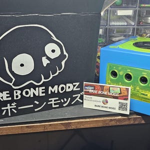 May include: A blue and green video game console with a black box featuring a skull graphic and the text "BARE BONE MODZ". A business card with the same logo is also present. The box has a "FRAGILE" sticker.