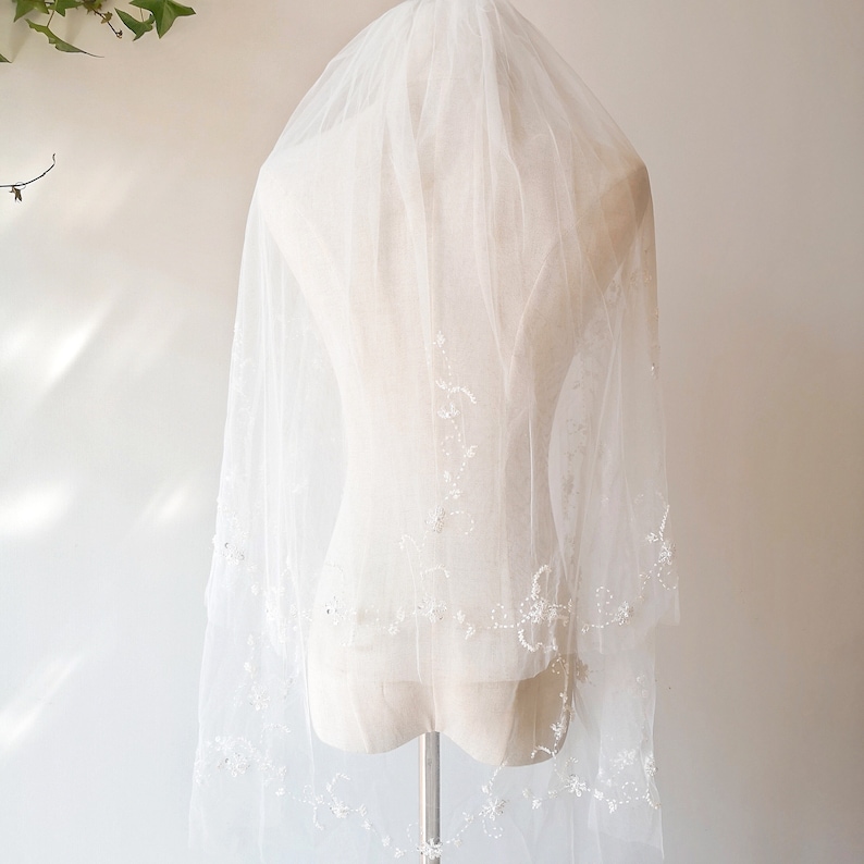 Pu&ograve; includere: Un velo da sposa bianco con ricami floreali delicati e perline. Il velo &egrave; esposto su un manichino bianco.