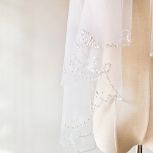 May include: A white bridal veil with a beaded trim. The veil is made of soft tulle and has a delicate, feminine look.