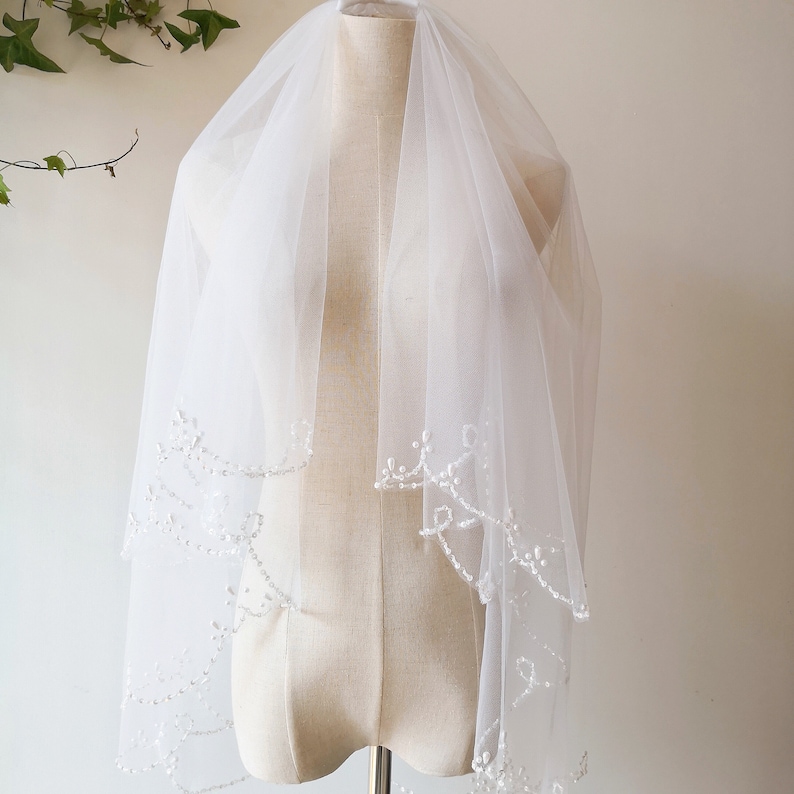 May include: A white bridal veil with a delicate beaded trim. The veil is layered and has a scalloped edge.