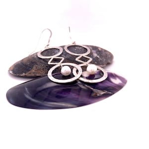 May include: A pair of silver earrings with a geometric design and a pearl dangle. The earrings are displayed on a purple seashell.